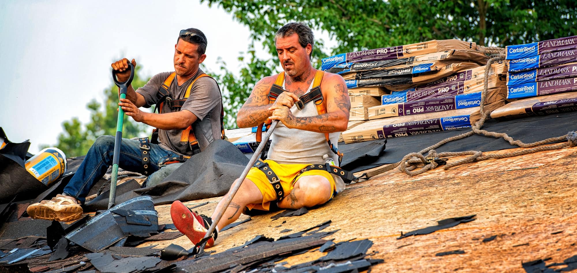 Two construction workers in safety gear repair a pitched roof, tearing off shingles with tools as bundles of roofing material sit nearby.