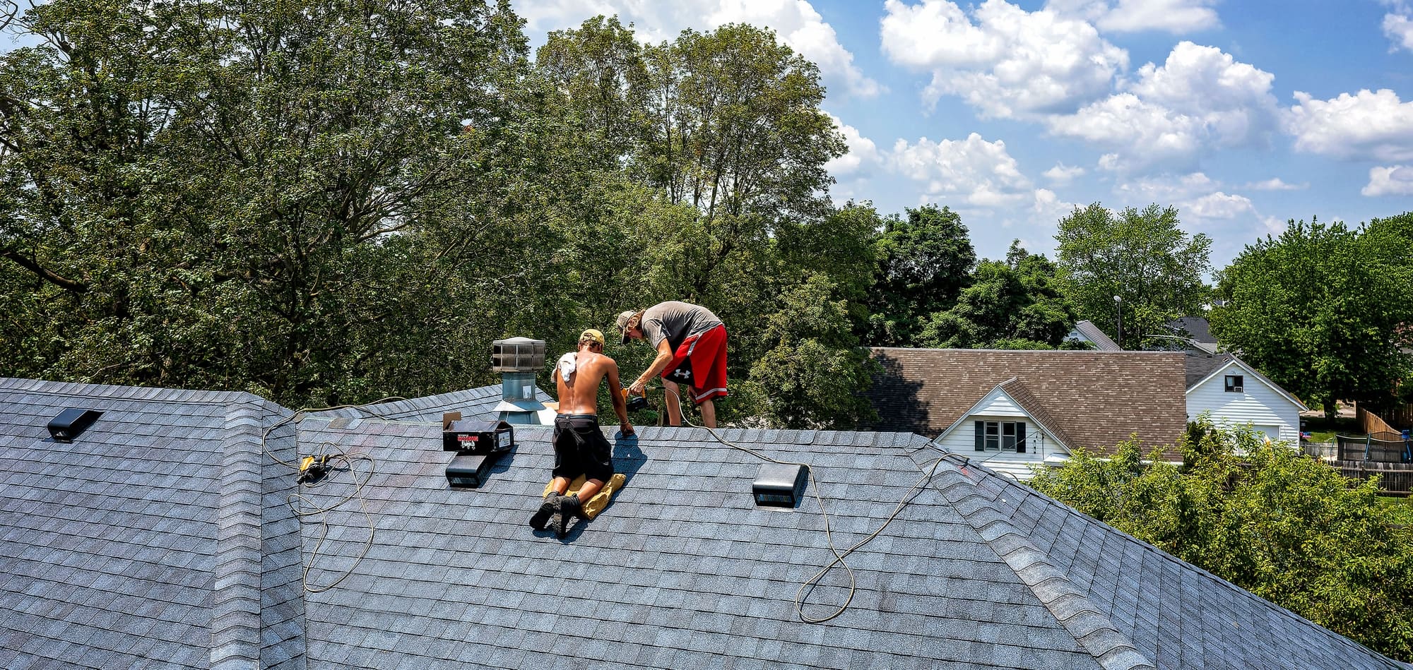 Two workers on a sloped roof wiring or installing equipment, ropes and tools around them, residential neighborhood in the background