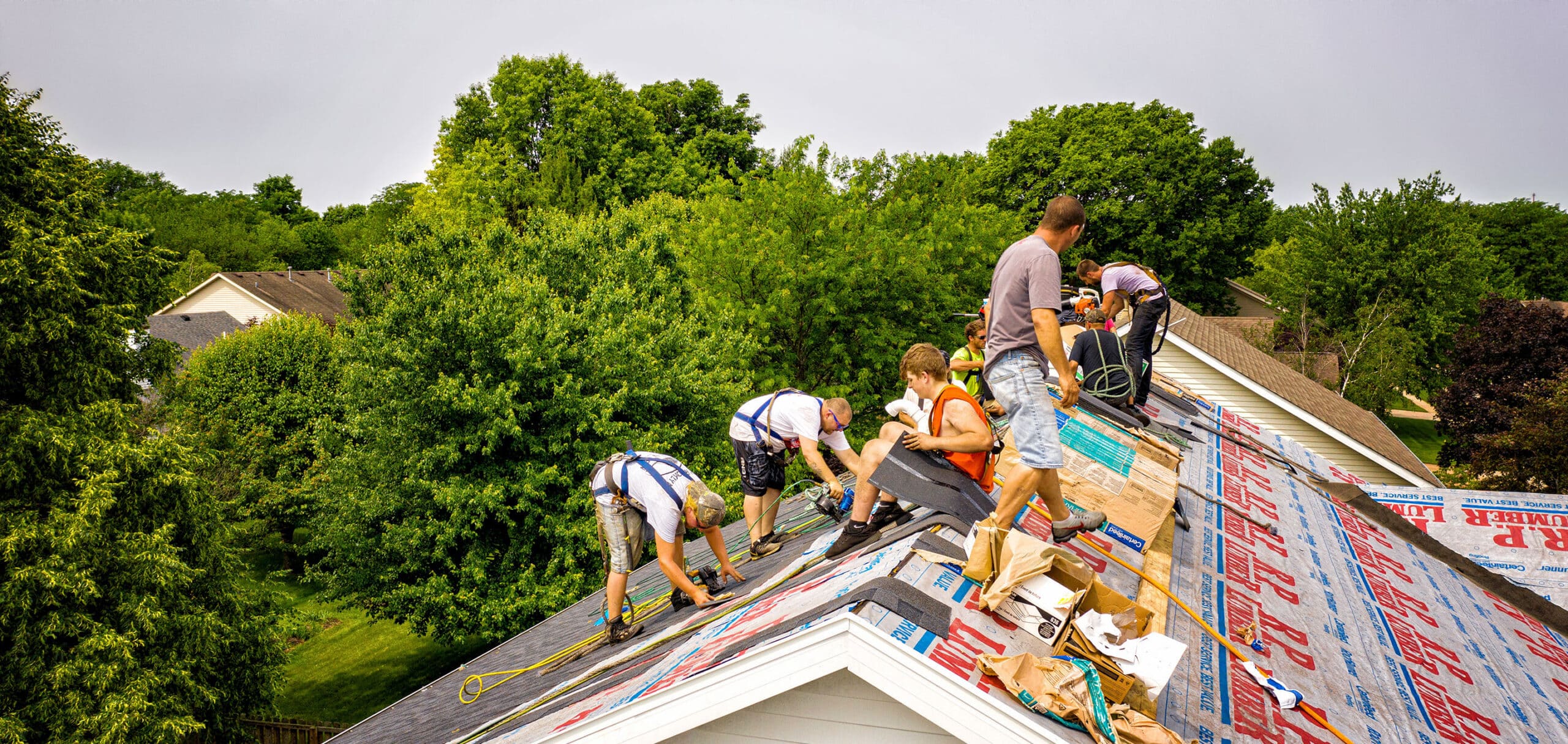 Construction workers on a sloped roof wearing harnesses and safety gear, laying shingles and organizing tools amid trees and houses in the background.
