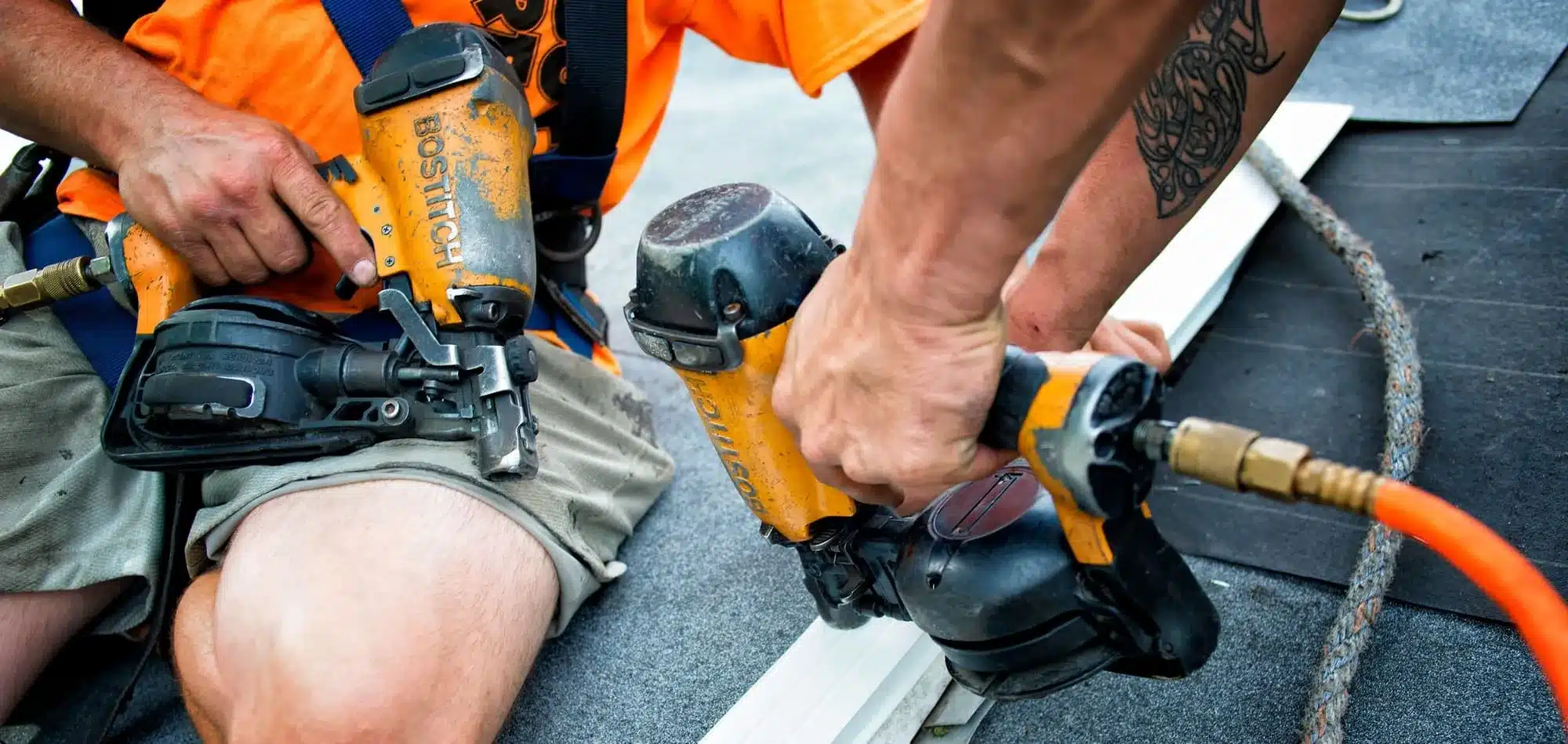 Construction worker in an orange shirt using a yellow pneumatic nail gun on a roof, with tools and rope nearby