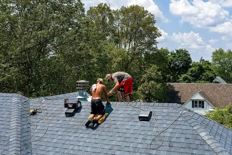 Two workers on a gray shingled residential roof repairing or inspecting it, with tools, a vent pipe, and cords nearby, trees and houses in the background.