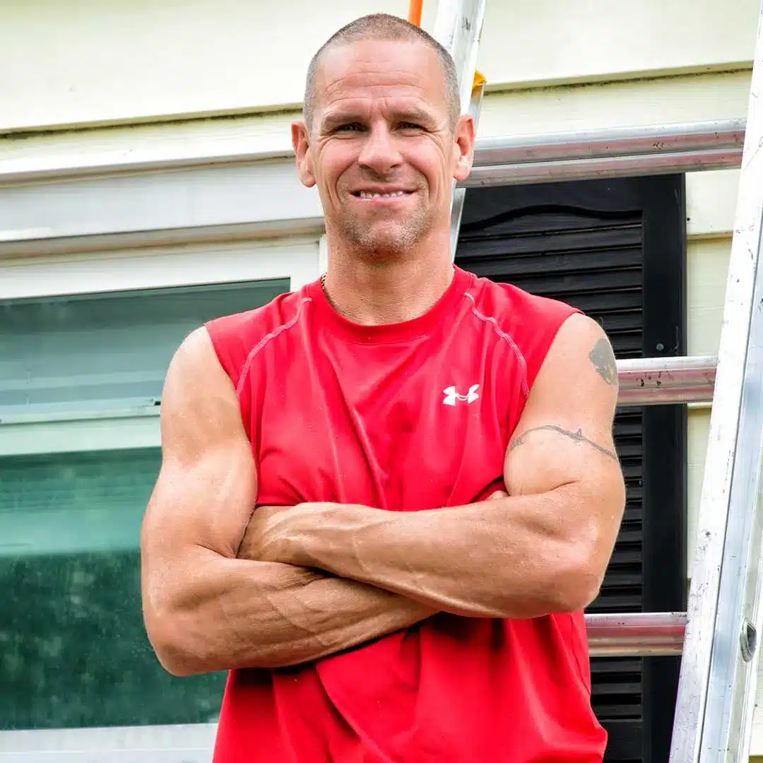 Muscular man in a red sleeveless shirt with arms crossed, standing beside a ladder outdoors.