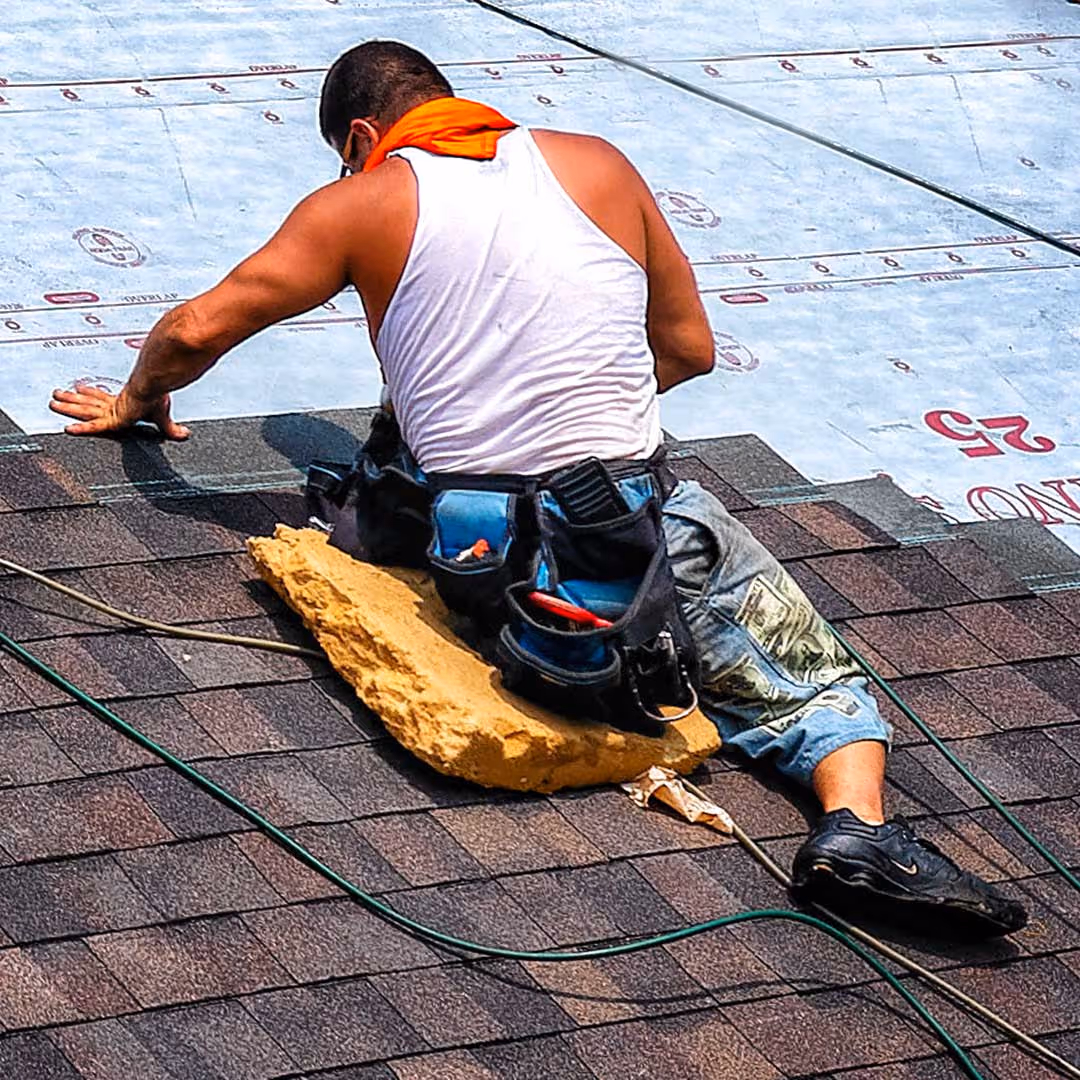 Roofing worker kneels on shingles, installing insulation/underlayment with tool belt and bright orange scarf.