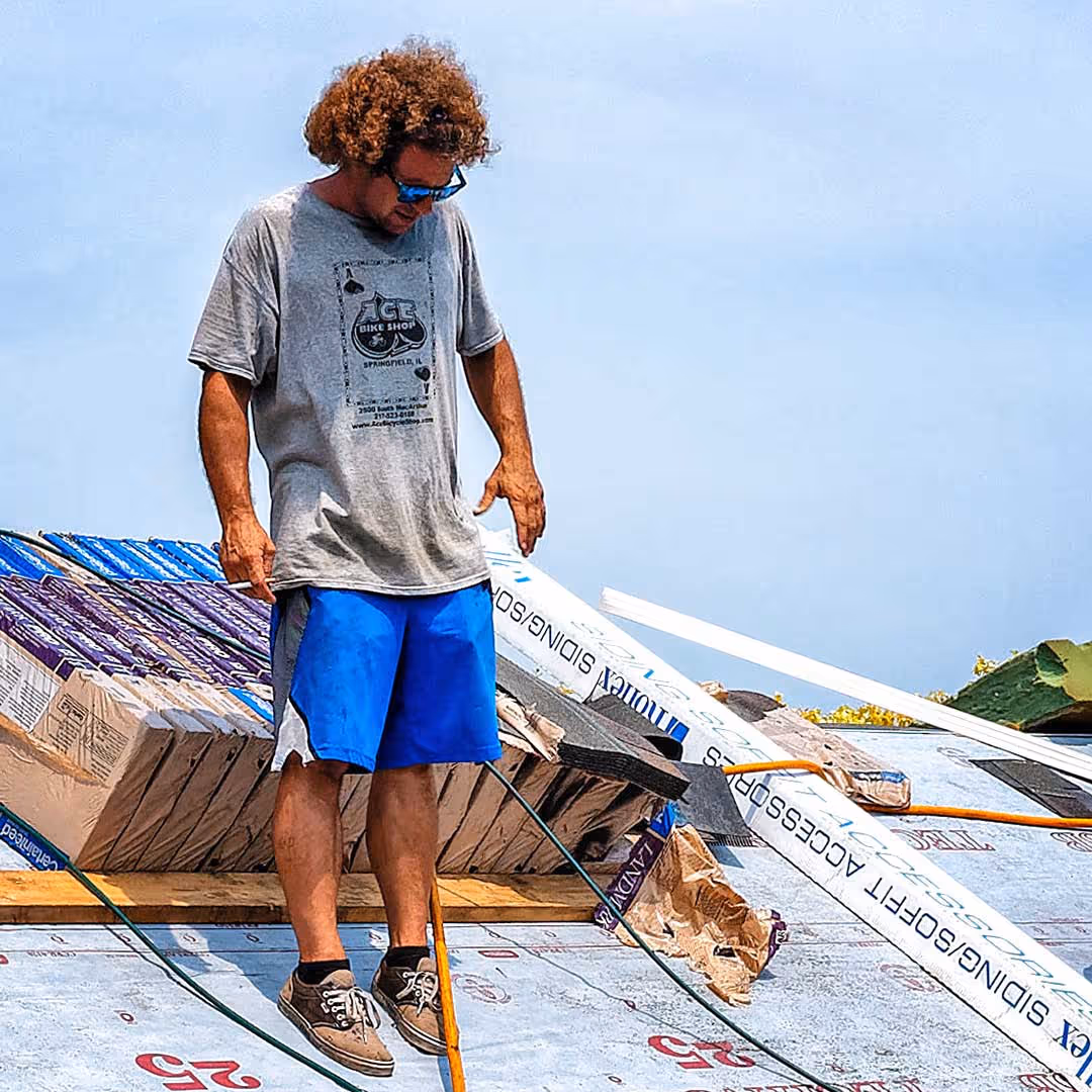 Man with curly hair in a gray t‑shirt and blue shorts stands on a partially built roof with insulation and roofing materials nearby under a blue sky.