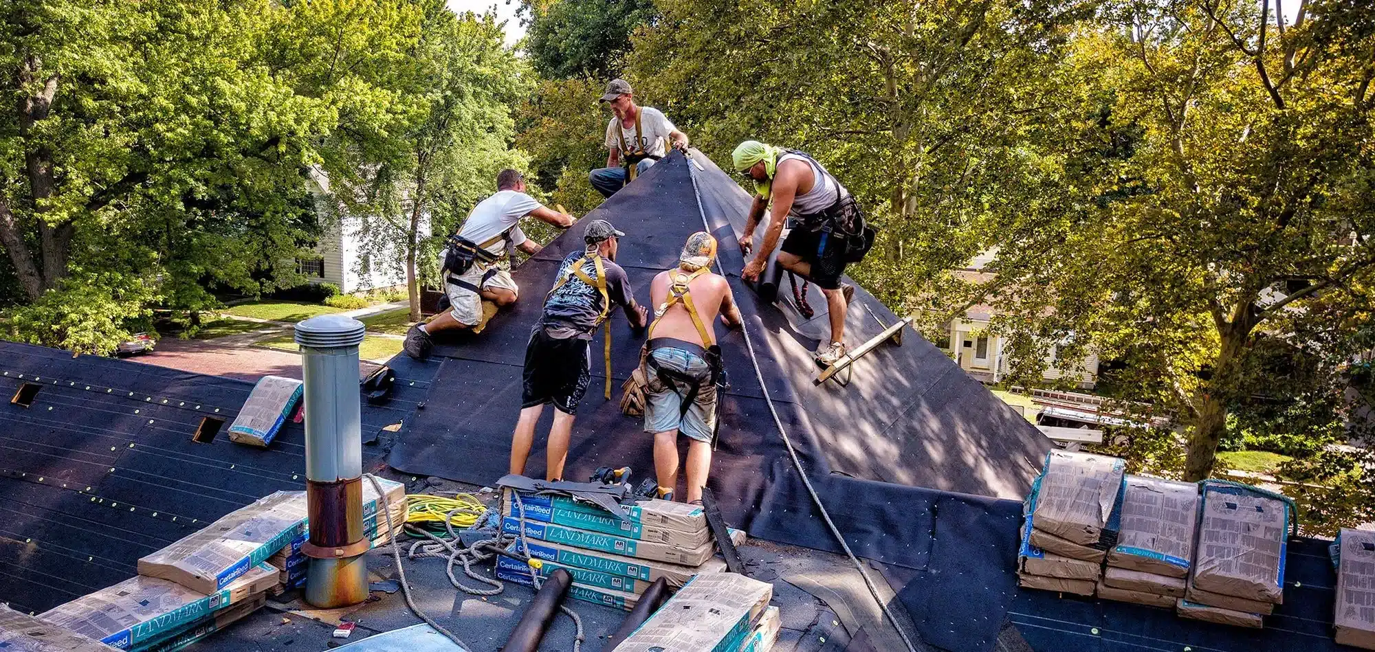 Five construction workers on a steep black shingled roof, installing underlayment with tools and safety harnesses nearby.