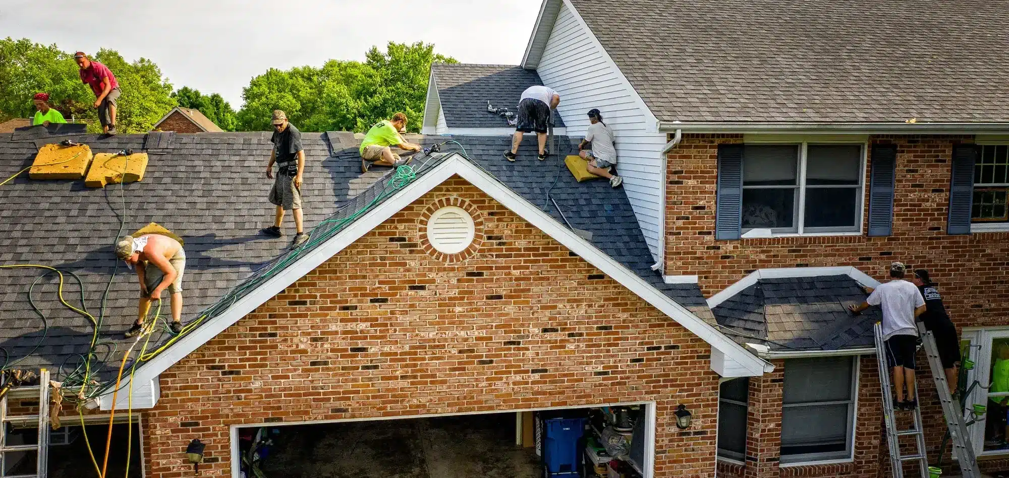Construction workers on a pitched roof replacing shingles, with hoses and tools scattered nearby and ladders at the house edge