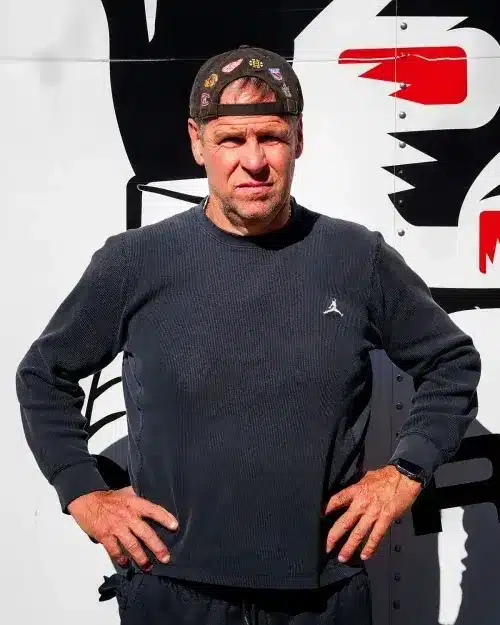 Man in a dark sweater with a backward-cap, hands on hips, posing in front of a graphic truck wall.