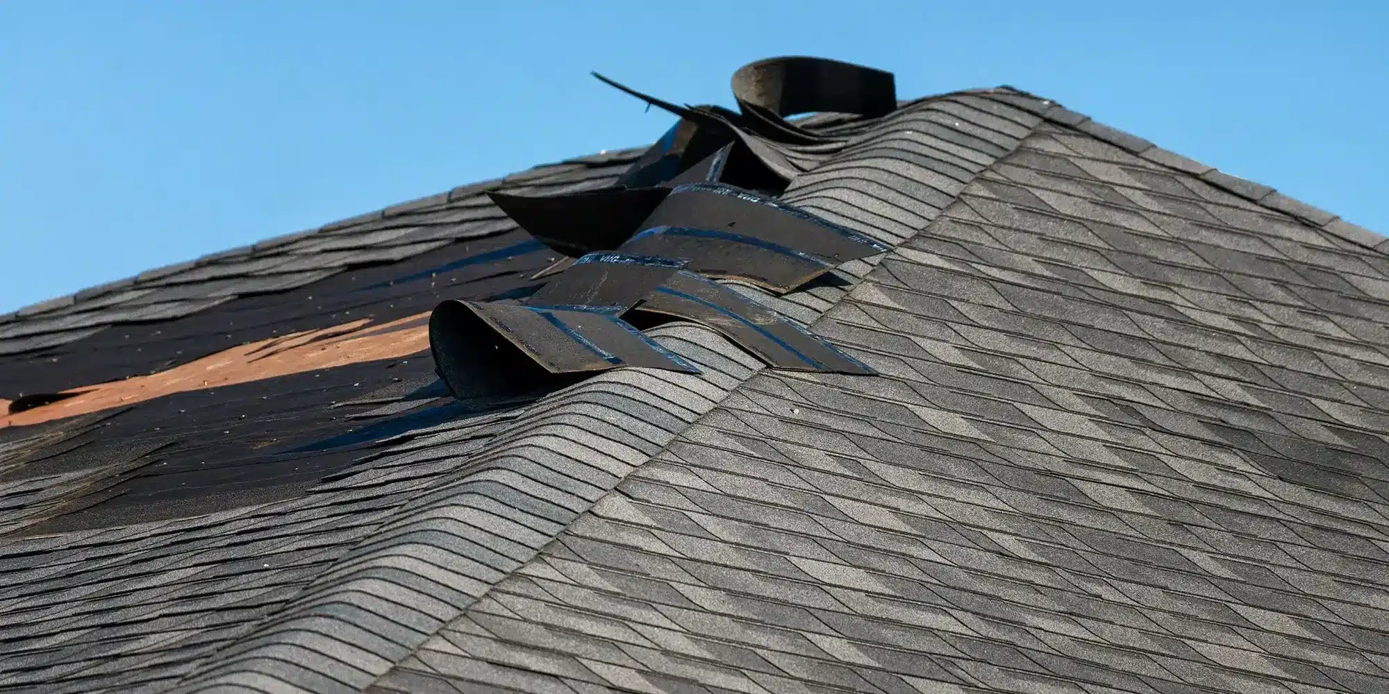 Pitched gray shingle roof with bent metal flashing strips curling upward against a clear blue sky