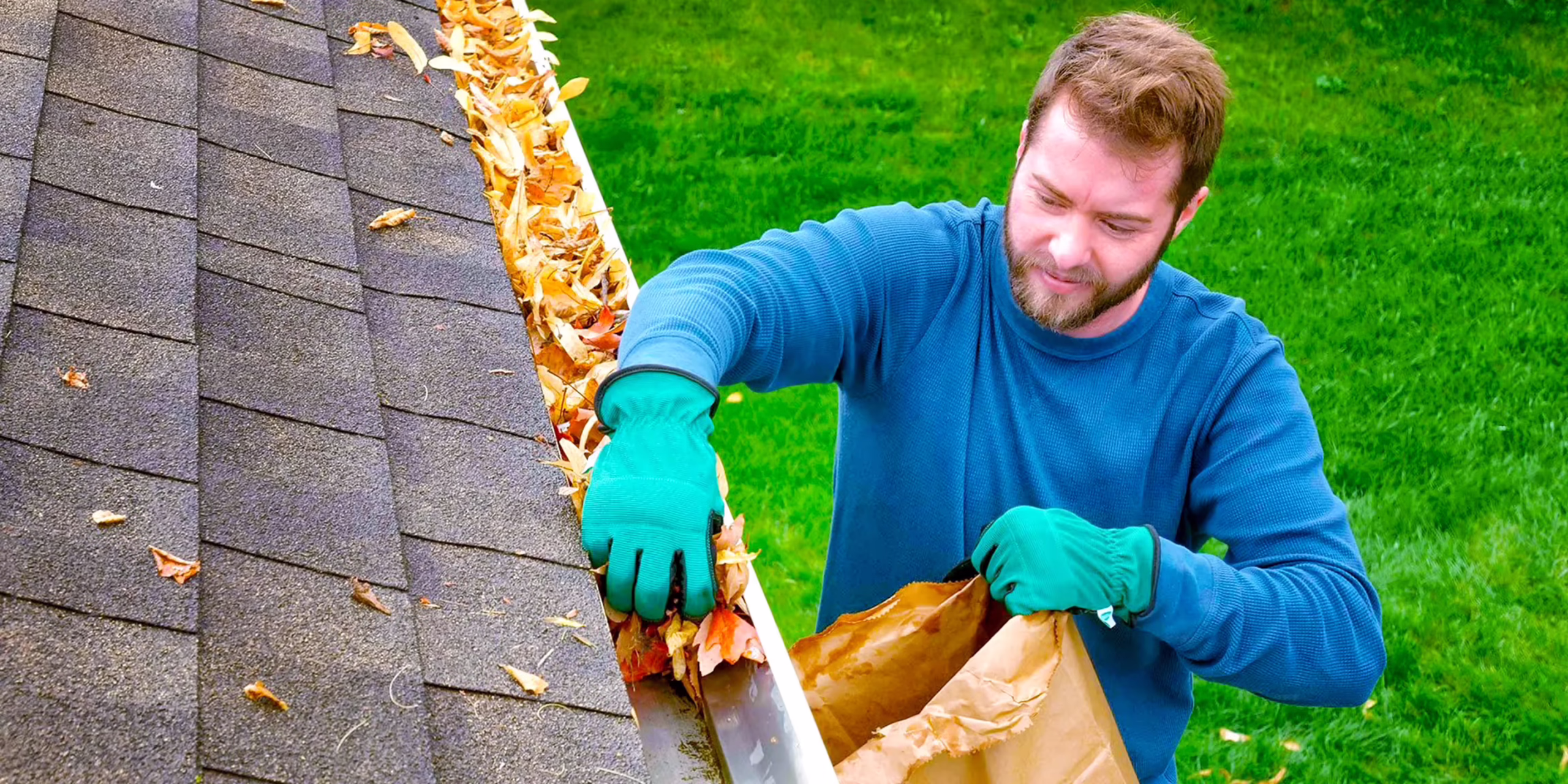 Man in a blue shirt and green gloves on a roof collecting fallen leaves into a paper bag. Green lawn in background.